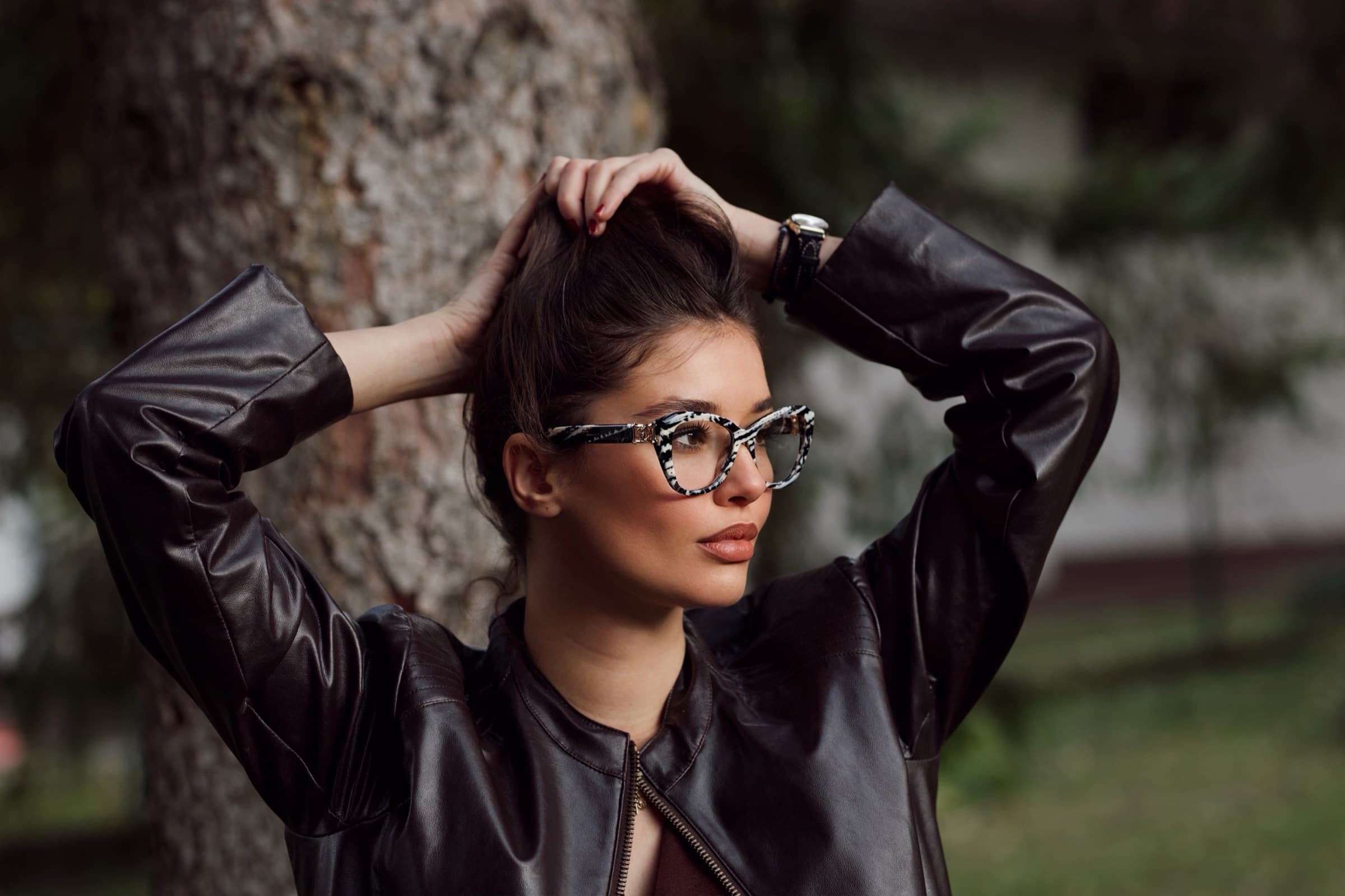 Hands in her hair, tortoiseshell frames, against a pine trunk
