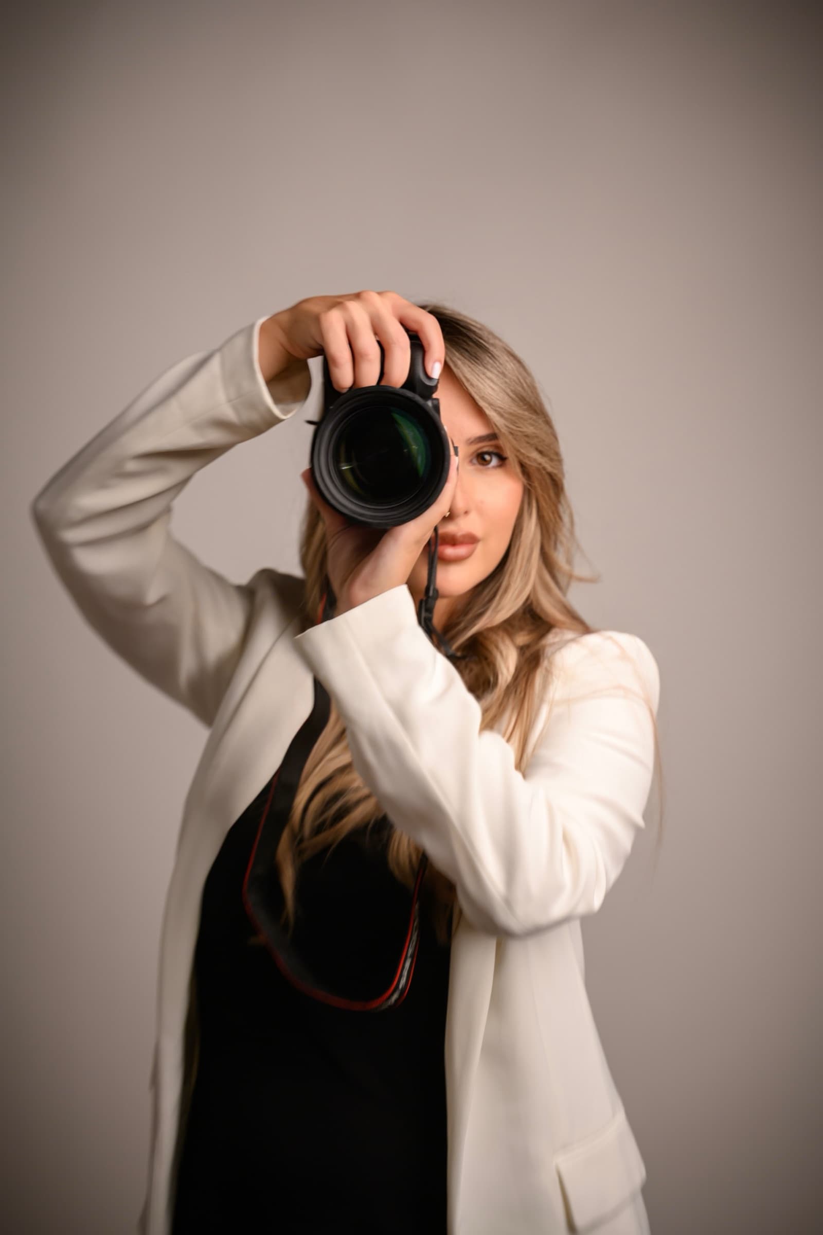Maja Topić raising her camera to the lens, in a white blazer