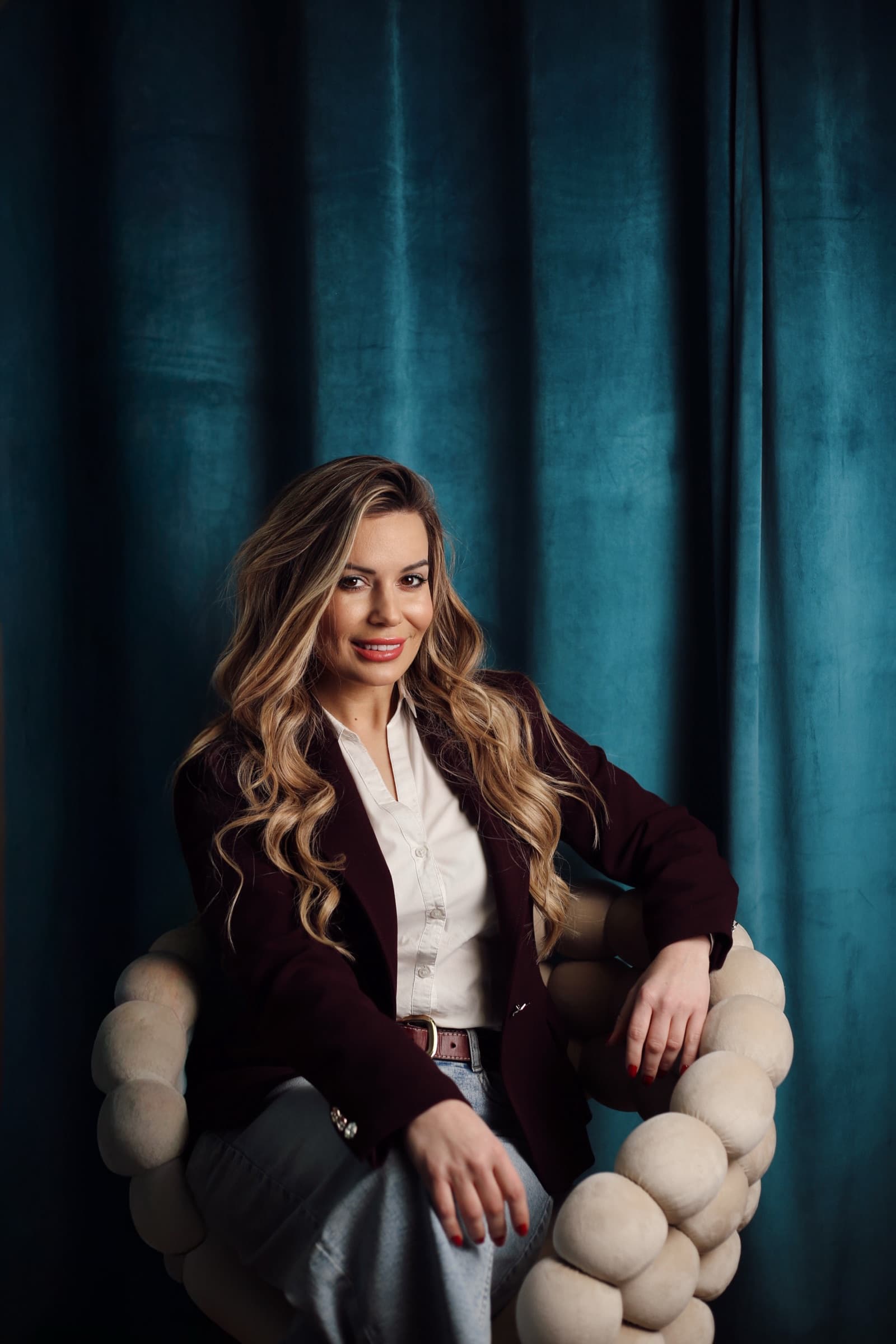 Seated against a teal velvet curtain in a burgundy blazer