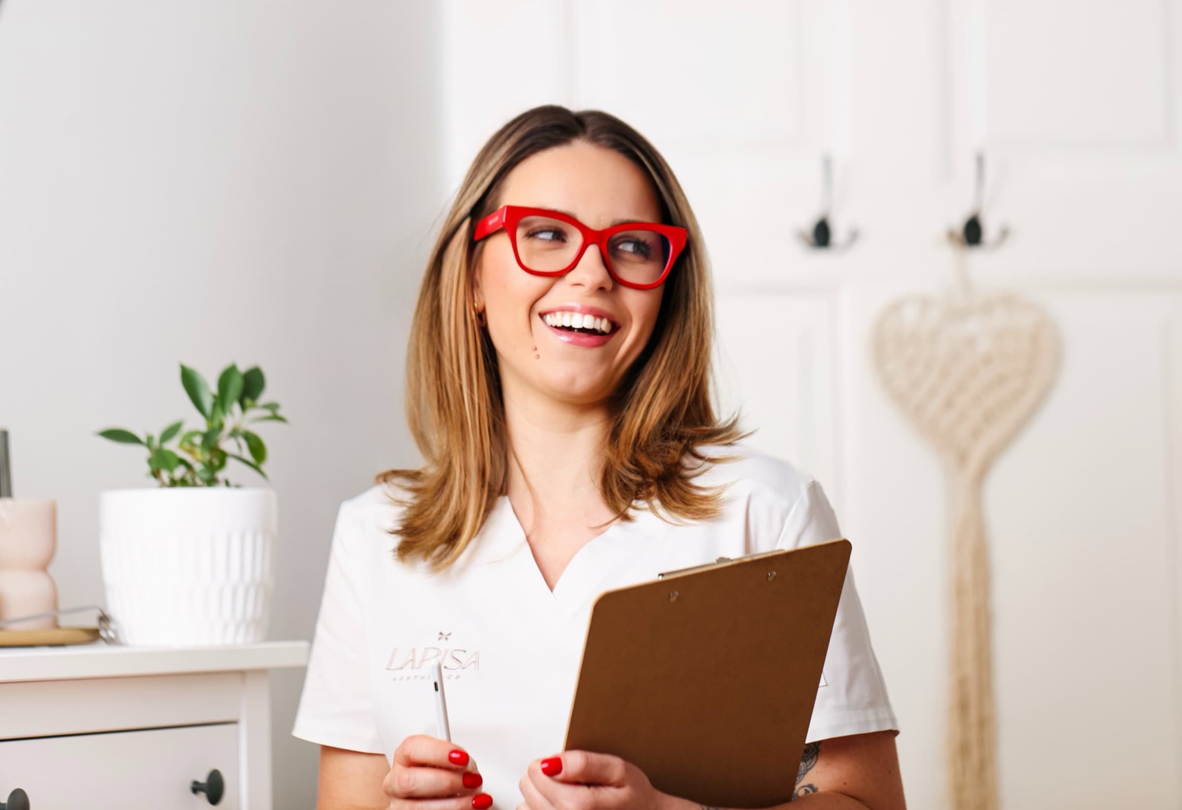 Aesthetics practitioner in branded scrubs and red glasses, laughing with a clipboard in her studio